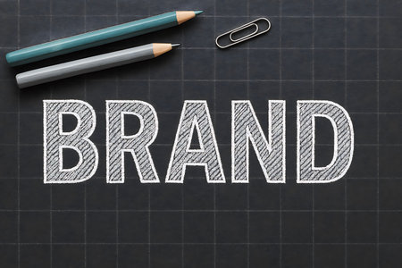 Word BRAND written in chalk on a dark chalkboard, accompanied by pencils and a paperclip, symbolizing marketing and business concepts.の素材