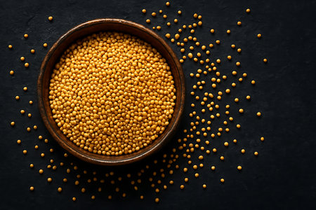 Yellow mustard seeds fill a rustic ceramic bowl, with additional seeds scattered across a textured dark surface. Capturing an aerial view of this common spice ingredient.の素材