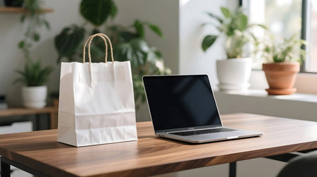 Clean wooden desk featuring a white paper shopping bag and an open laptop, set against a blurred background of vibrant green plantsの素材