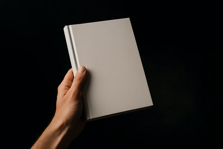 A hand holds a closed blank beige hardcover book against a dark and moody, shadowed backdropの素材