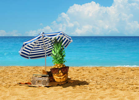 Happy pineapple character having summer holiday under blue-white umbrella on hot sandy beach by turquoise blue oceanの写真素材