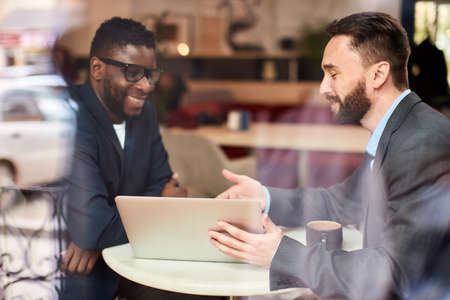 Two businessmen causacian and african american having a conversation in cafe in front of laptop, through the windowの写真素材