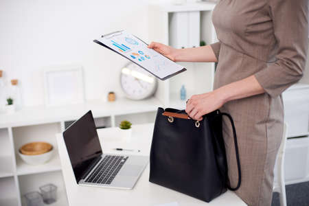 Businesswoman in brown dress holds a notepad above black leather bag, which stands on the white table, laptop stands nearby.の写真素材