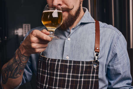Tattooed man tastes beer from the glass he holds in a close up.の写真素材