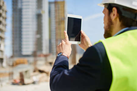 An engineer in white helmet and green safety vest works on his tablet at the construction spot.の写真素材