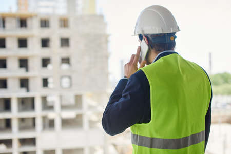 An engineer in a helmet and green safety vest talks on the phone.