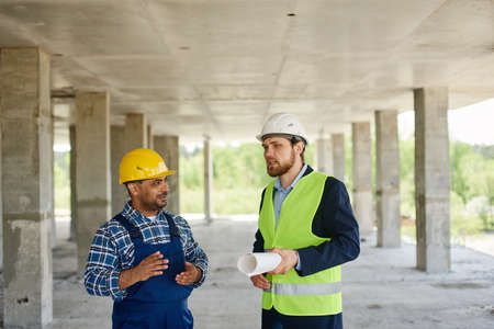Two engineers discuss work related questions together holding folded blueprint.