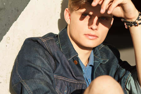 Stylish man in denim jacket sitting near wall of buildingの写真素材