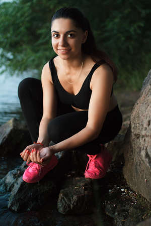 Pretty sportswoman portrait sitting on a large stone near river. Evening.の写真素材