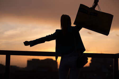 Good buy. Silhouette of a girl with bag on the roof at sunset in the cityの写真素材