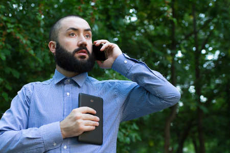 Man with beard hold tablet computer and speak into mobile in summer park
