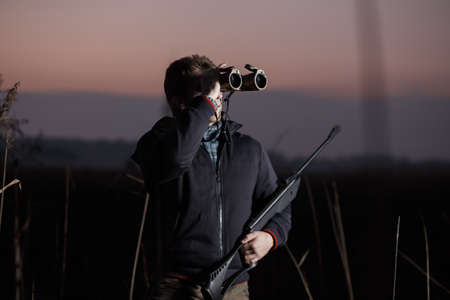 Hunter man with rifle see in binocular in field in eveningの写真素材