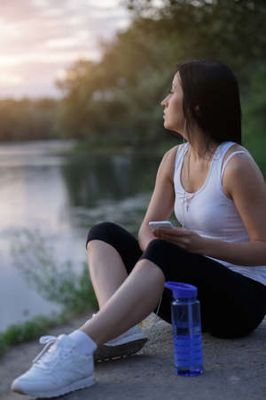 Woman runner listen music after workout and looking at sunset near pondの写真素材