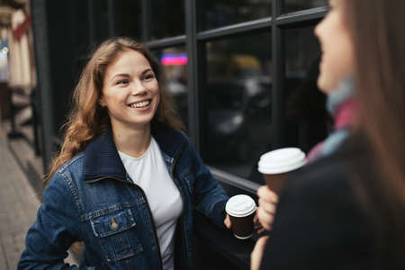 Two young women are happy to drink coffee in city. They holding a paper cups and smiling.