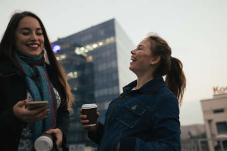 Outdoors portrait of two cheerful girls drinking coffee. Walking in a city.の写真素材