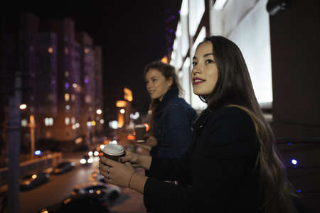 Two young woman friends looking at city street from balcony at nightの写真素材