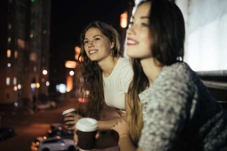 Two young woman friends looking at city street from balcony at nightの写真素材