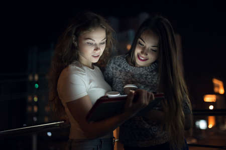Two woman friends look down on digital tablet in balcony at nightの写真素材
