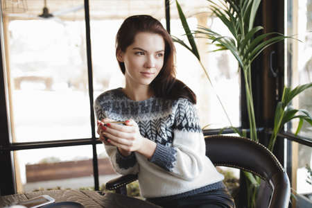 Woman in pullover sitting with cup of coffee in a cafe and look at windowの写真素材