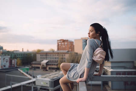 Stylish young woman sitting on roof and relaxation, evening city backgroundの写真素材