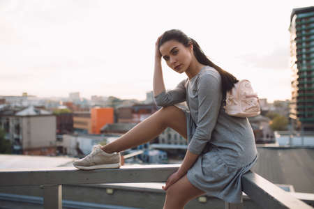 Stylish young woman sitting on roof and posing, evening city backgroundの写真素材