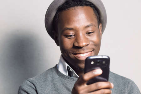 Portrait of smiling african american man in hat using mobile phone near grey brick wallの写真素材