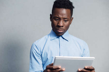 African man embarrassed by what he saw on the screen of the tablet PC at grey wall background