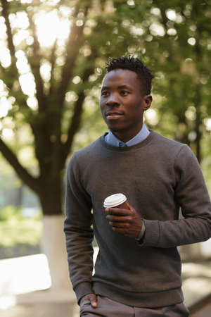 African american man walking and drinking tea from paper cup in sunny evening city streetの写真素材