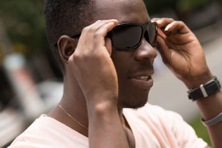 Portrait of handsome african american man sitting in city on bench, he wearing sunglasses on green tree background and looking upの写真素材