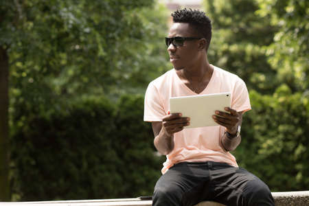 Portrait of stylish fashionable african american man in sunglasses sitting at city park on bench and using his tablet PC, green tree backgroundの写真素材