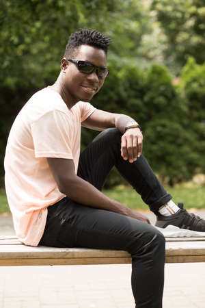 Portrait of stylish fashionable african american man in sunglasses sitting at city park on bench, green tree backgroundの写真素材