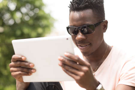 Portrait of stylish fashionable african american man in sunglasses sitting at city park on bench and using his tablet PC, green tree background, closeup shotの写真素材