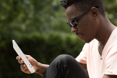 Portrait of stylish fashionable african american man in sunglasses sitting at city park on bench and using his tablet PC, green tree background, closeup shotの写真素材