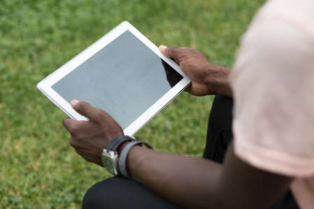 African american male hands holding tablet pc on grass background. Black man using touchpad in a park.の写真素材