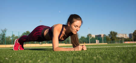 Young happy sportswoman in sportswear making plank exercise on stadium field area outdoors. Healthy...