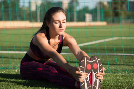 Young woman in sportswear stretches her leg sitting on stadium grass outdoors. Healthy lifestyle concept. Workout sport activity.の写真素材