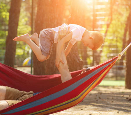 Father holding son above him lying in hammock, they resting at pine forest, recreation conceptの写真素材