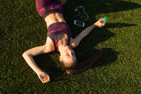 Young tired sportswoman in sportswear lying and relaxing after hard workout training on stadium green grass outdoors. Healthy lifestyle concept. Summer sport activity.の写真素材