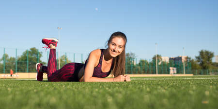 Happy young smiling sportswoman in sportswear lying and relaxing on stadium field area outdoors. Healthy lifestyle concept, sport activity.の写真素材