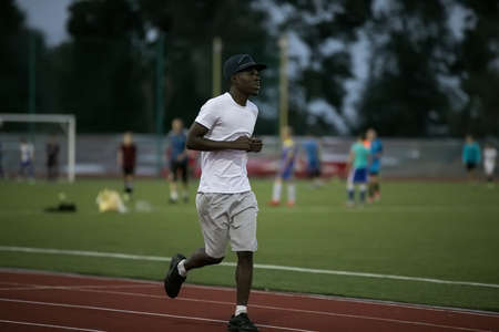 African male sportsman in a cap running on stadium track, he trains endurance