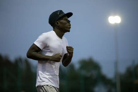 African male sportsman in a cap running on stadium track, he trains enduranceの写真素材