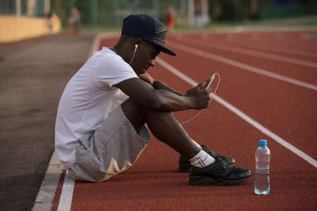 African american male sportsman sit on stadium track and listen music after hard workoutの写真素材