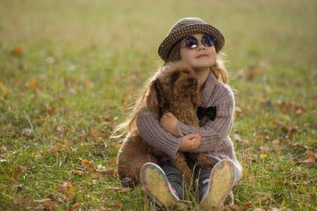 Little girl in hat sitting on grass hugging dog together at the autumn meadow, art portrait