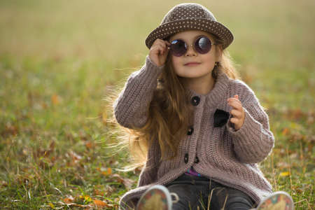 Little girl with sunglasses and hat sitting on a grass in autumn clothesの写真素材