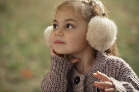 Portrait of cute little girl in autumn clothes and headphones which warming her ears, sitting on grass at city parkの写真素材