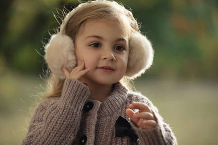Portrait of cute little girl in autumn clothes and headphones which warming her ears, sitting on grass at city parkの写真素材