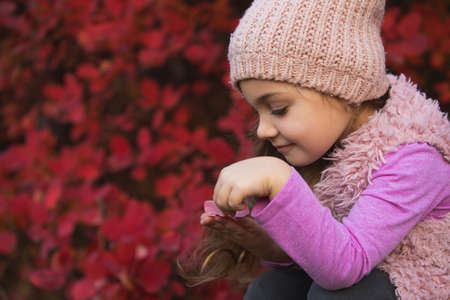 Little girl portrait in hat at autumn clothes against red tree foliage background, she plays with...