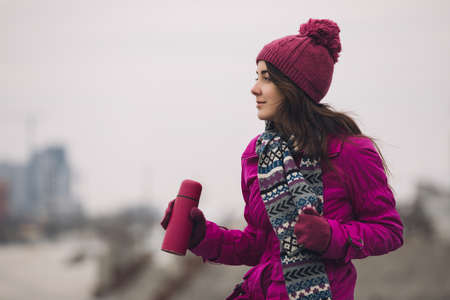 Woman in bright winter jacket holding thermos with tea at city backgroundの写真素材