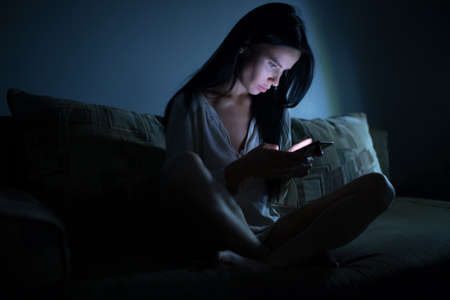 Tired woman using blue smartphone screen at night dark roomの写真素材