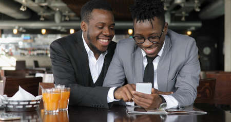 African man friends laughing in cafe, looking photos together on smart phoneの写真素材